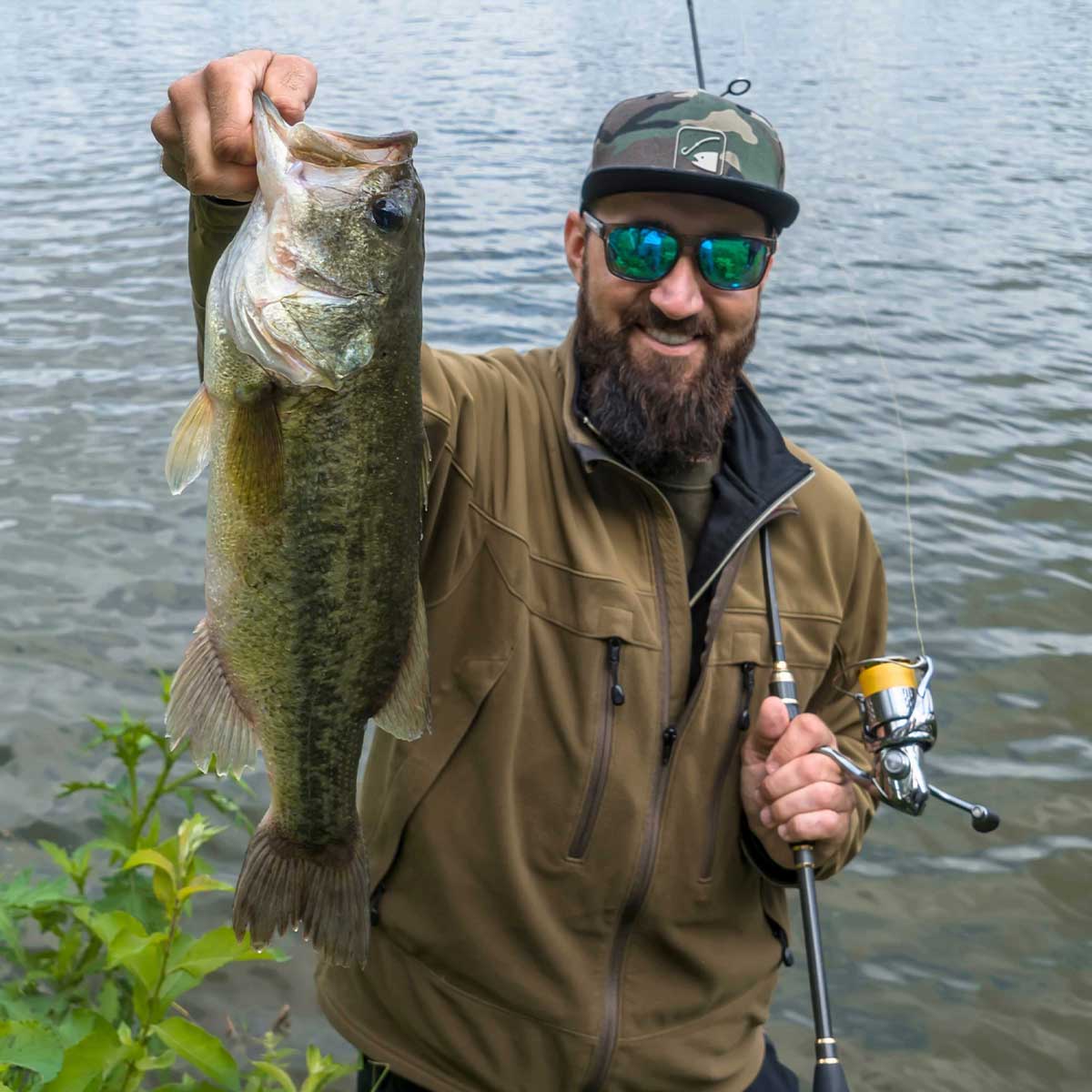 A man wearing sunglasses and a cap smiles while holding up a large fish in one hand and a fishing rod in the other, standing by the waters edge.