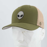 The Eternal Glyph Alien Gray (Double Layer Silicone) Flexfit® Mesh Trucker Cap features an olive green and beige design with a black and white alien face logo, shown on a white geometric mannequin—perfect for UFO pop culture fans.