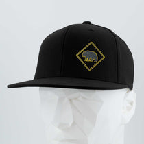The Eternal Glyph Bear Crossing Sign (Silicone) - Flexfit® Flat Bill Cap features a black snapback with a yellow diamond bear patch, shown on a mannequin head against a plain background, evoking the wilderness spirit.