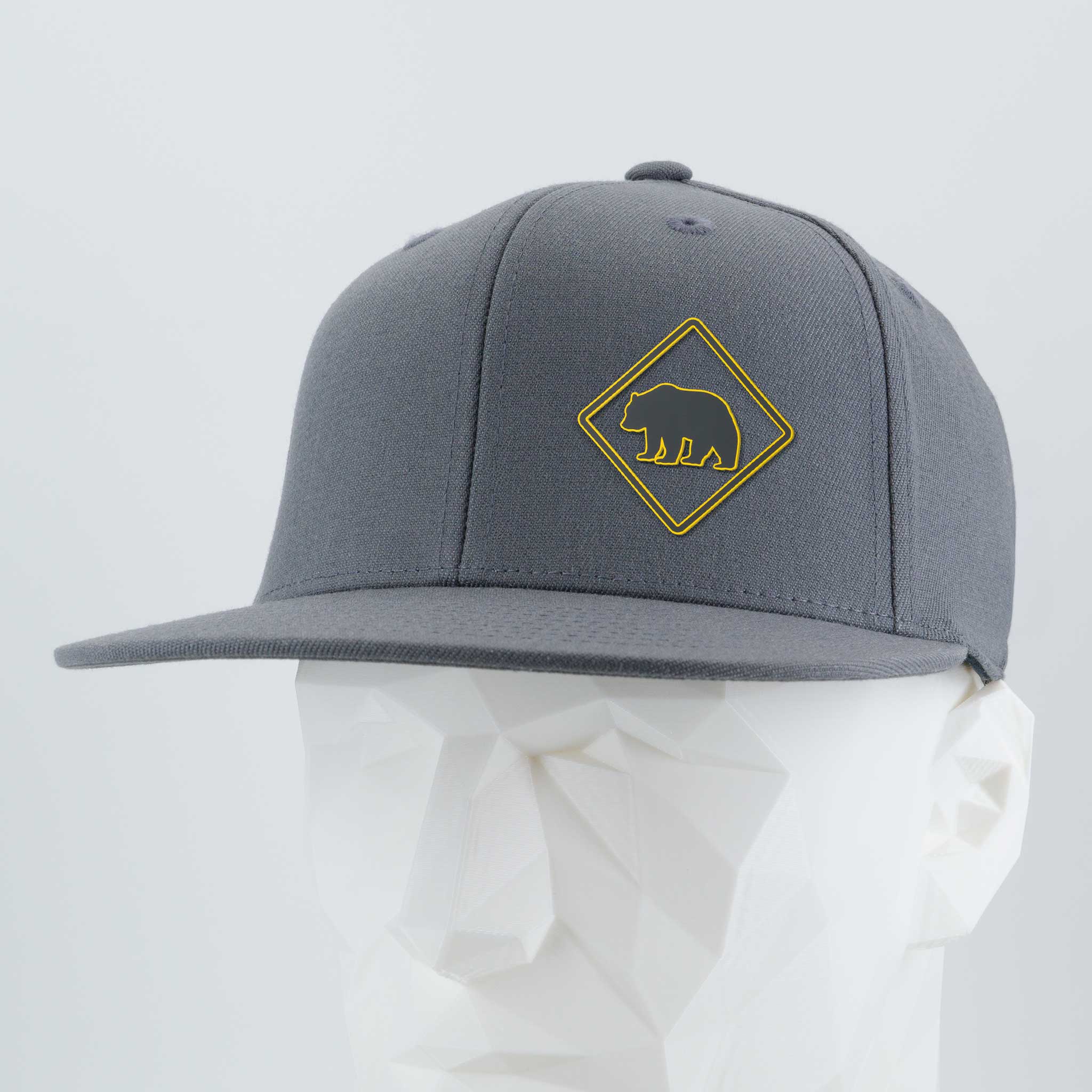 The Eternal Glyph Bear Crossing Sign (Silicone) Flexfit® Flat Bill Cap features a yellow diamond bear patch on gray, shown on a white geometric mannequin.