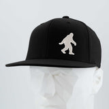The Eternal Glyph Bigfoot Silhouette Albino (Silicone) Flexfit® Flat Bill Premium Fitted Cap in black features a bold Bigfoot silhouette on the front, presented on a geometric white mannequin head against a neutral background.