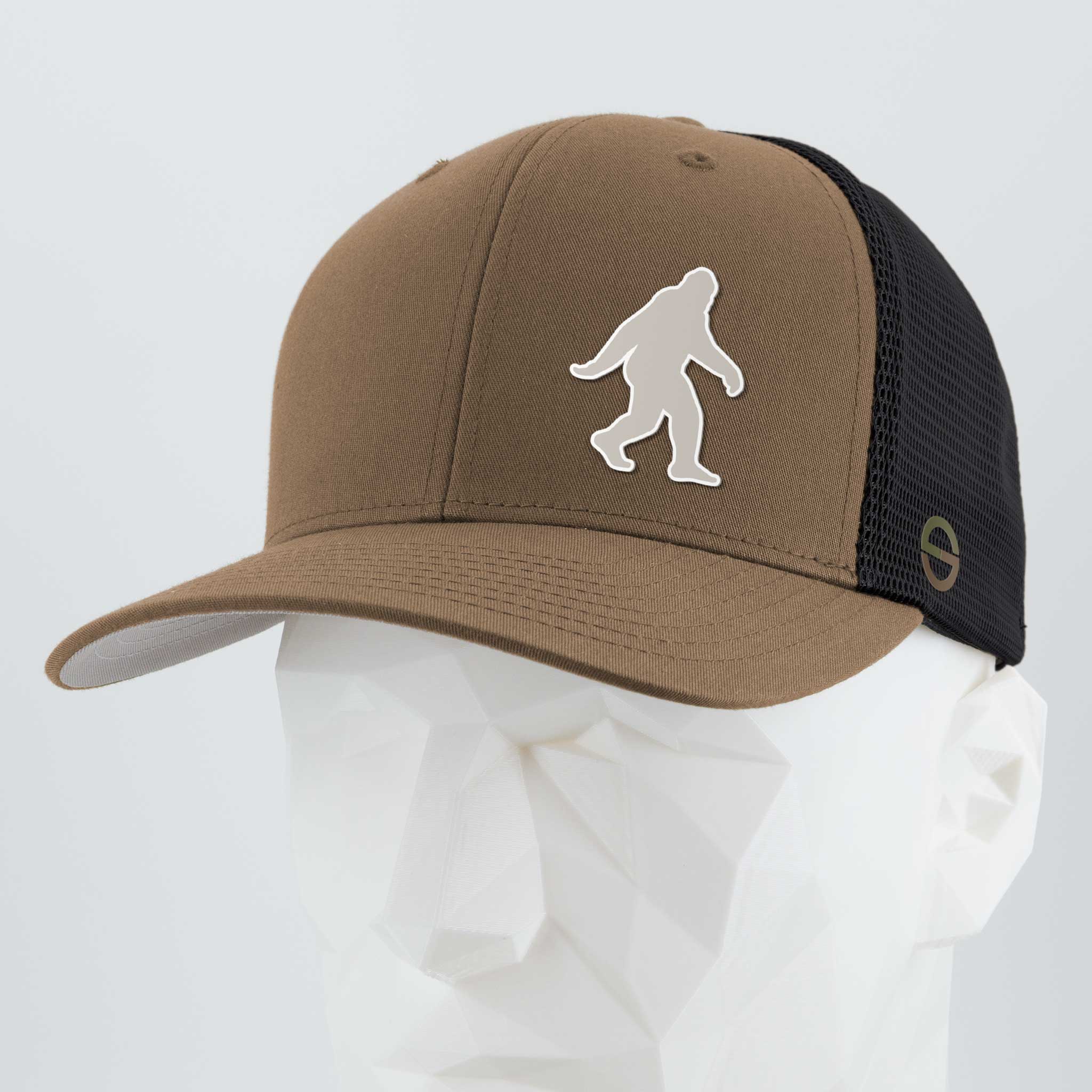 The Eternal Glyph Bigfoot Silhouette Albino (Silicone) Flexfit® Mesh Trucker Cap in brown and black features a white Bigfoot silhouette, inspired by Pacific Northwest folklore, shown on a faceted white mannequin head against a plain background.