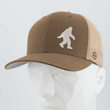 The Eternal Glyph Bigfoot Silhouette Albino Flexfit® Mesh Trucker Cap features a brown and beige design with a white Bigfoot logo, inspired by Pacific Northwest folklore, shown on a faceted white mannequin head.