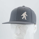 The Eternal Glyph Bigfoot Silhouette Albino (Silicone) Flexfit® Flat Bill Premium Fitted Cap in gray features a bold Bigfoot design on the front, bringing wilderness folklore style to this modern, flat-brim hat. Displayed on a white mannequin head.