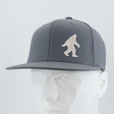 The Eternal Glyph Bigfoot Silhouette Albino (Silicone) Flexfit® Flat Bill Premium Fitted Cap in gray features a bold Bigfoot design on the front, bringing wilderness folklore style to this modern, flat-brim hat. Displayed on a white mannequin head.