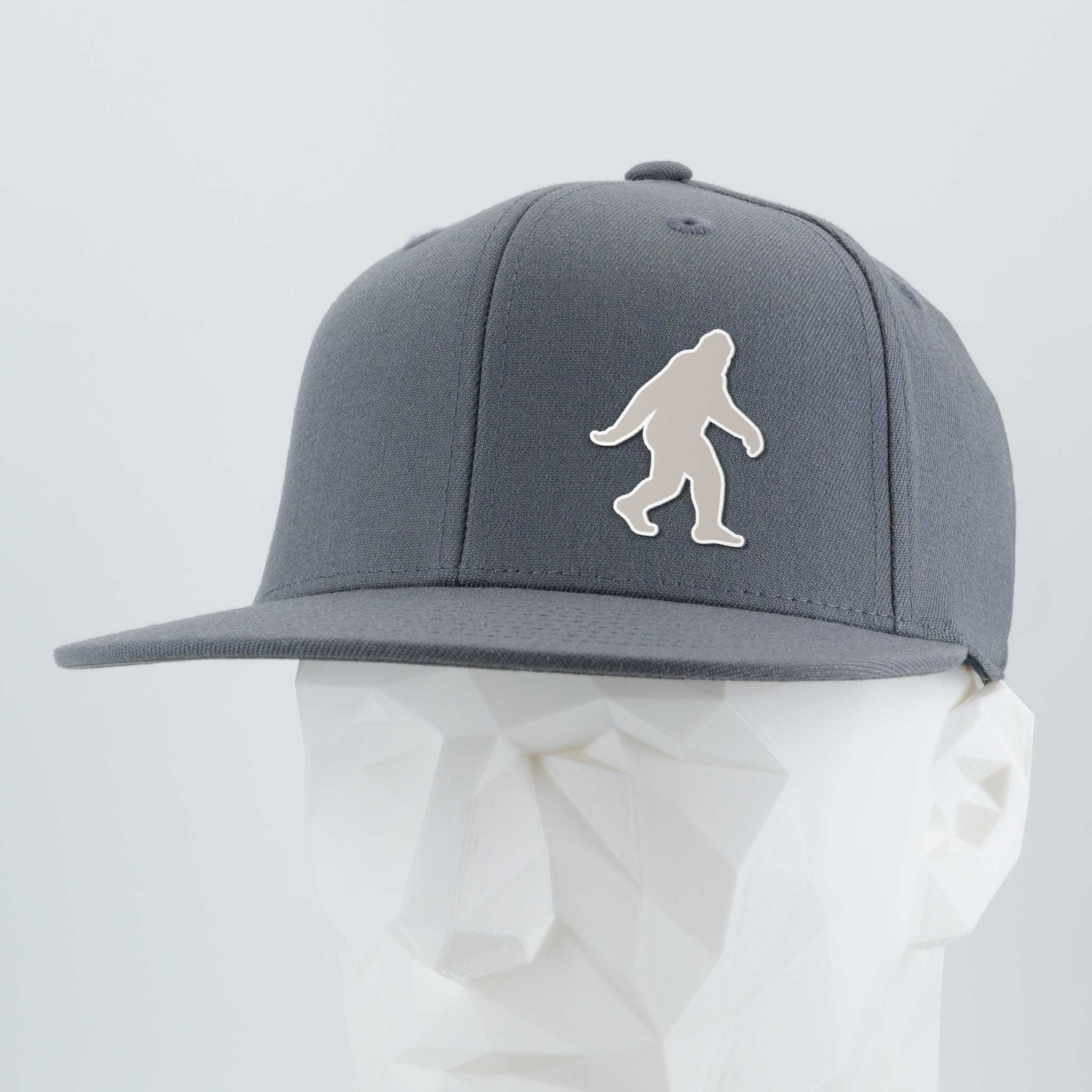 The Eternal Glyph Bigfoot Silhouette Albino (Silicone) Flexfit® Flat Bill Premium Fitted Cap in gray features a bold Bigfoot design on the front, bringing wilderness folklore style to this modern, flat-brim hat. Displayed on a white mannequin head.