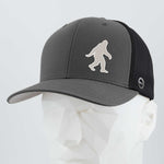 The Eternal Glyph Bigfoot Silhouette Albino Flexfit® Mesh Trucker Cap features a white Sasquatch logo on dark gray and black, inspired by Pacific Northwest folklore, shown on a white geometric mannequin head.