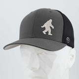 The Eternal Glyph Bigfoot Silhouette Albino Flexfit® Mesh Trucker Cap features a white Sasquatch logo on dark gray and black, inspired by Pacific Northwest folklore, shown on a white geometric mannequin head.