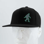 The Eternal Glyph Bigfoot Silhouette (Silicone) Flexfit® Flat Bill Premium Fitted Cap in black features a green Bigfoot design, shown on a white geometric mannequin head against a light gray background—perfect for outdoor lovers.