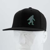 The Eternal Glyph Bigfoot Silhouette (Silicone) Flexfit® Flat Bill Premium Fitted Cap in black features a green Bigfoot design, shown on a white geometric mannequin head against a light gray background—perfect for outdoor lovers.