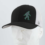 The Eternal Glyph Bigfoot Silhouette (Silicone) Flexfit® Mesh Trucker Cap features a green outlined Bigfoot icon on a black and white cap, shown here on a white geometric mannequin head.