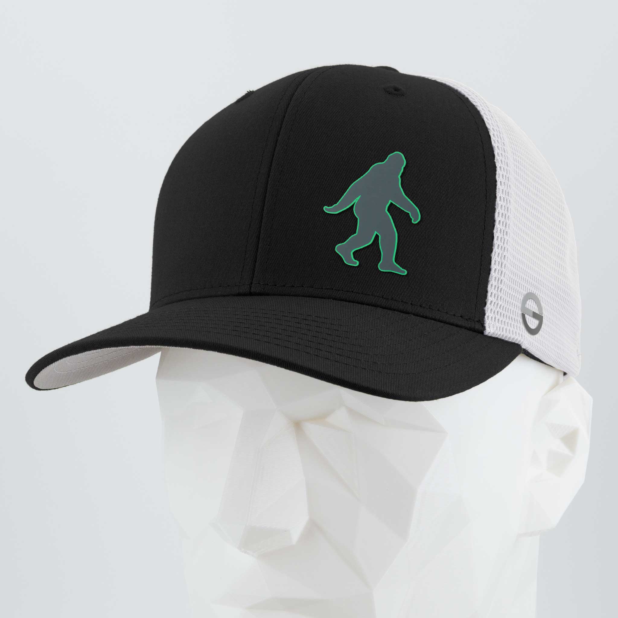 The Eternal Glyph Bigfoot Silhouette (Silicone) Flexfit® Mesh Trucker Cap features a green outlined Bigfoot icon on a black and white cap, shown here on a white geometric mannequin head.