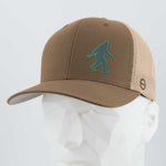 The Eternal Glyph Bigfoot Silhouette (Silicone) Flexfit® Mesh Trucker Cap in brown and beige, featuring a green Bigfoot icon, is shown on a white geometric mannequin head.