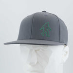 The Eternal Glyph Bigfoot Silhouette (Silicone) Flexfit® Flat Bill Premium Fitted Cap in gray features a green Bigfoot icon on the front, shown on a white geometric mannequin head against a light background.