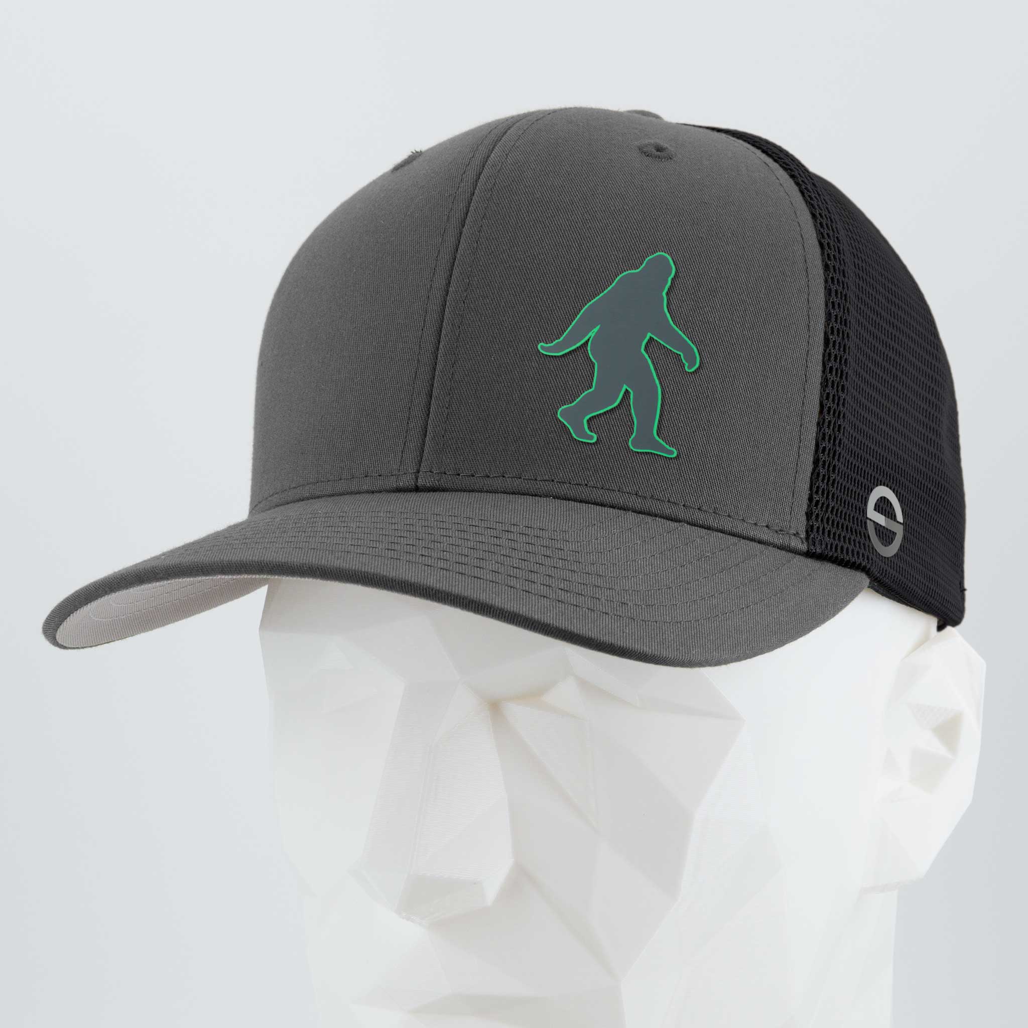 The Eternal Glyph Bigfoot Silhouette (Silicone) Flexfit® Mesh Trucker Cap features a gray and black mesh back and a green Bigfoot icon, modeled on a white mannequin—perfect for outdoor enthusiasts.