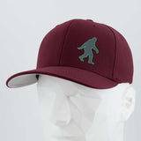 The Eternal Glyph Bigfoot Silhouette (Silicone) Flexfit® Premium Wool Blend Cap, in maroon with a green Bigfoot motif, is shown on a white geometric mannequin head against a light gray backdrop—ideal for outdoor enthusiasts.