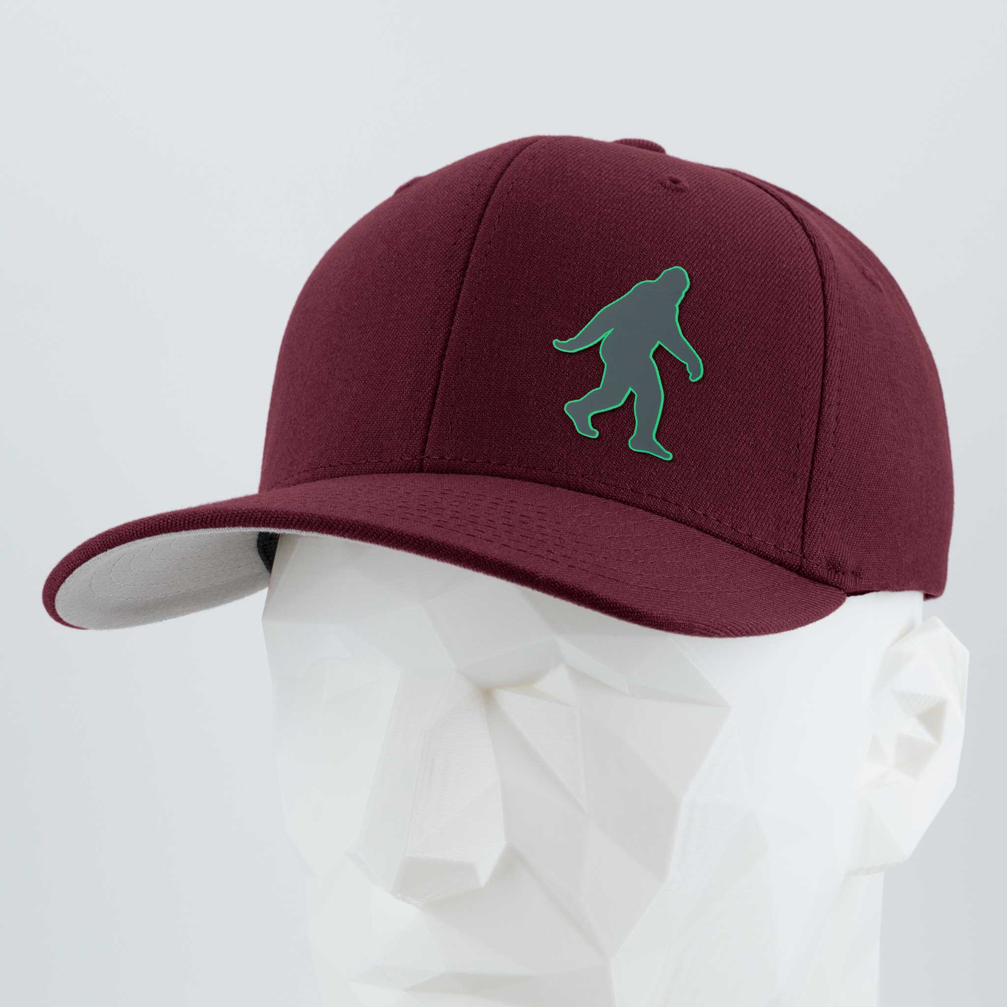 The Eternal Glyph Bigfoot Silhouette (Silicone) Flexfit® Premium Wool Blend Cap, in maroon with a green Bigfoot motif, is shown on a white geometric mannequin head against a light gray backdrop—ideal for outdoor enthusiasts.
