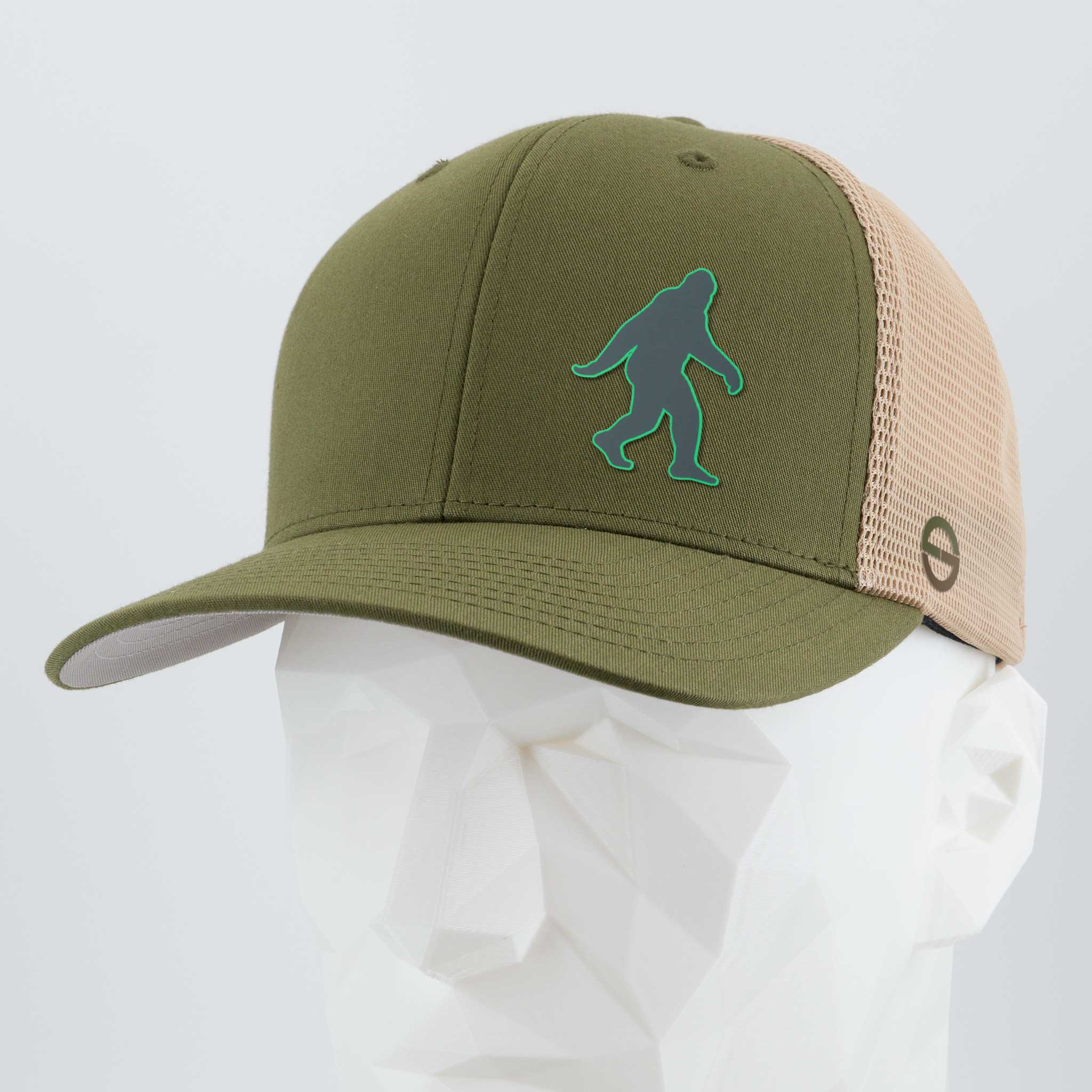 The Eternal Glyph Bigfoot Silhouette (Silicone) Flexfit® Mesh Trucker Cap in olive green and beige displays a blue-green Bigfoot adventure icon on the front, shown on a white geometric mannequin head.