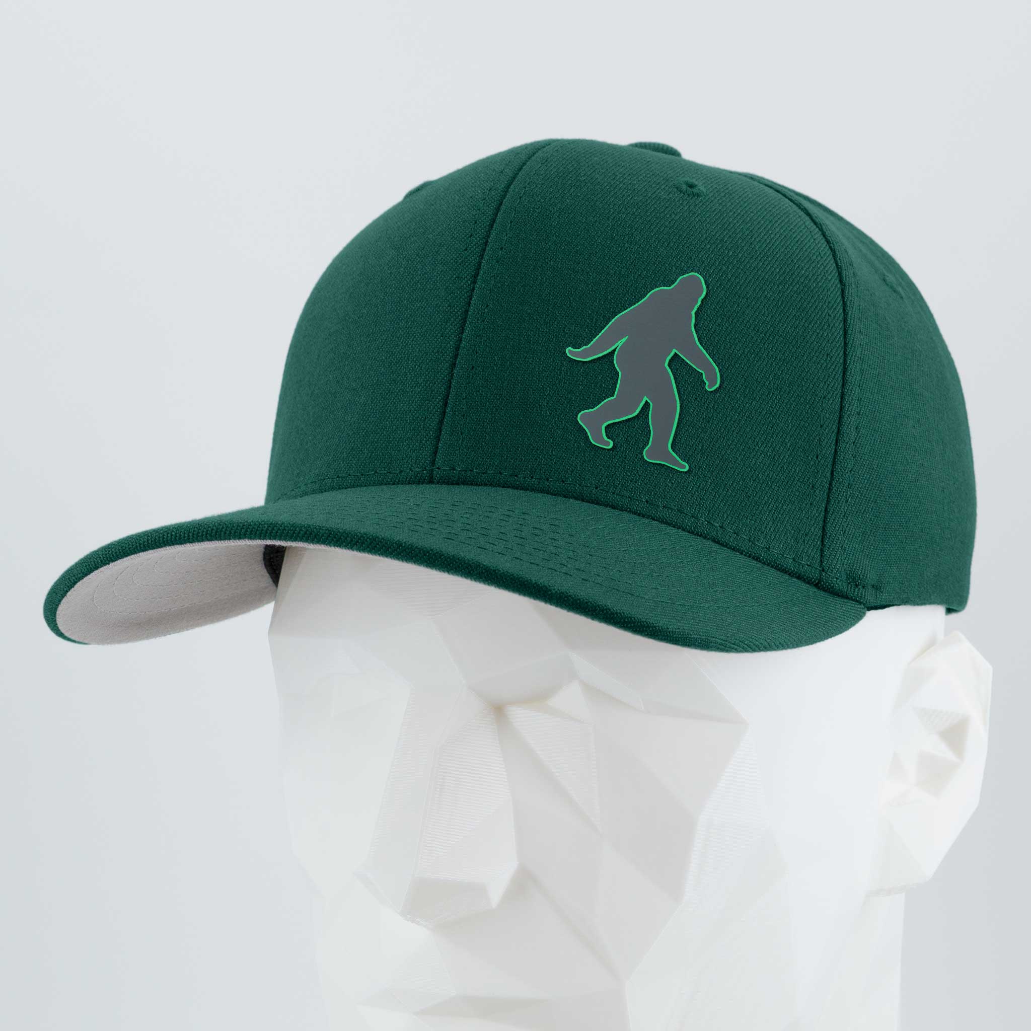 The Eternal Glyph Bigfoot Silhouette (Silicone) Flexfit® Premium Wool Blend Cap features a green and gray embroidered Bigfoot on the front, shown on a white mannequin head—ideal for any adventure lover or wilderness fan.