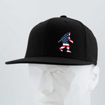The Eternal Glyph Patriotic Bigfoot (Silicone) Flexfit® Flat Bill Premium Fitted Cap in black features a silicone American flag Bigfoot silhouette, displayed on a white geometric mannequin against a light background.