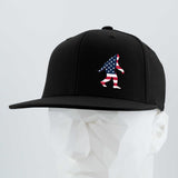 The Eternal Glyph Patriotic Bigfoot (Silicone) Flexfit® Flat Bill Premium Fitted Cap in black features a silicone American flag Bigfoot silhouette, displayed on a white geometric mannequin against a light background.