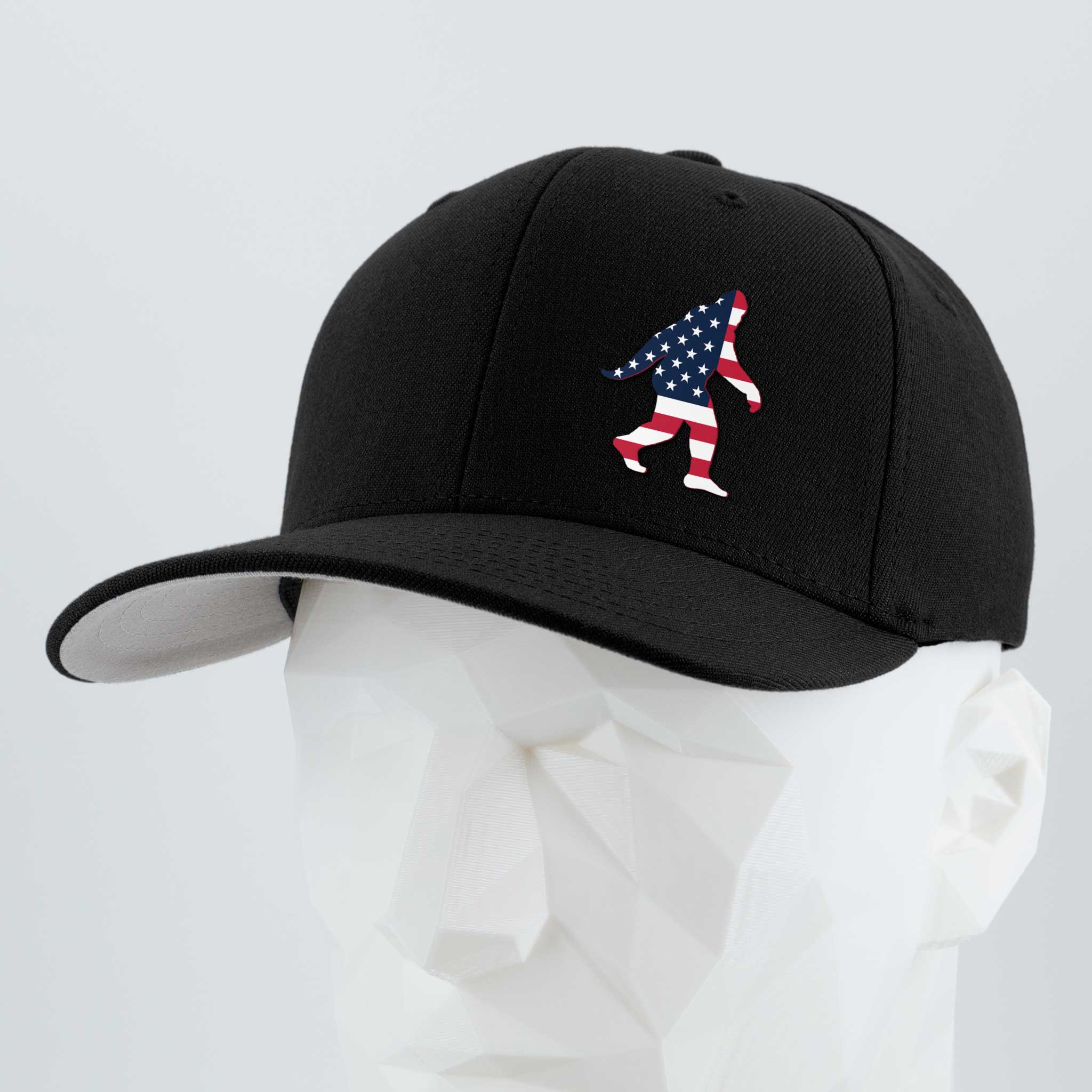 The Eternal Glyph Patriotic Bigfoot (Silicone) Flexfit® Premium Wool Blend Cap in black features a small American flag Sasquatch silhouette on the front, shown on a white geometric mannequin head against a plain background.