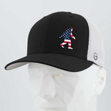 The Eternal Glyph Patriotic Bigfoot (Silicone) Flexfit® Mesh Trucker Cap features a black and white design with a silicone Bigfoot in an American flag motif—ideal for fans of wilderness folklore. Displayed on a white geometric mannequin head.