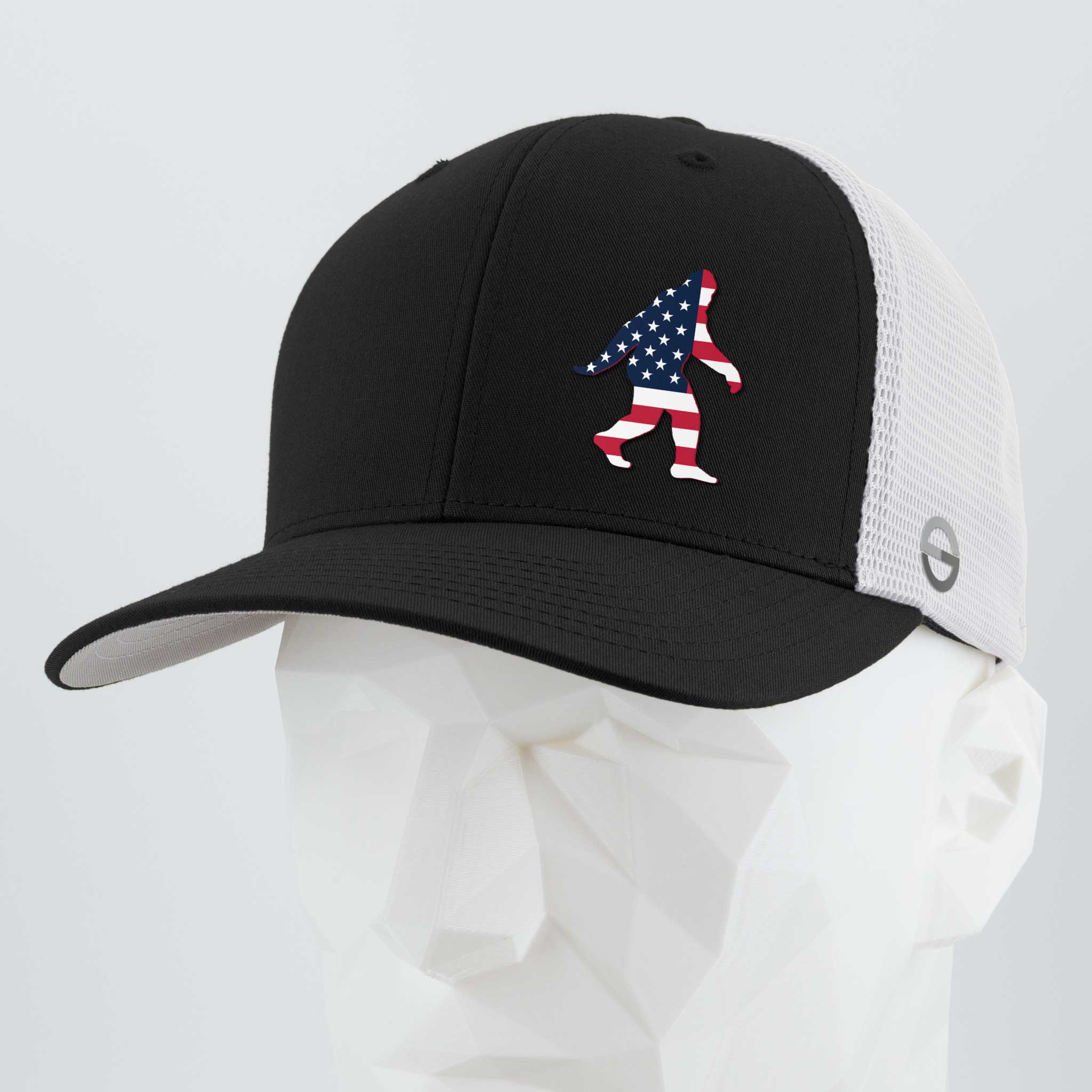The Eternal Glyph Patriotic Bigfoot (Silicone) Flexfit® Mesh Trucker Cap features a black and white design with a silicone Bigfoot in an American flag motif—ideal for fans of wilderness folklore. Displayed on a white geometric mannequin head.