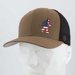 The Eternal Glyph Patriotic Bigfoot (Silicone) Flexfit® Mesh Trucker Cap is a brown and black hat with a silicone Patriotic Bigfoot patch in an American flag pattern, shown on a faceted white mannequin head.