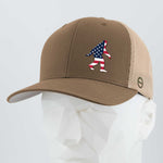 The Eternal Glyph Patriotic Bigfoot (Silicone) Flexfit® Mesh Trucker Cap features a tan and beige design with an American flag-patterned Bigfoot silhouette, displayed here on a white mannequin head.
