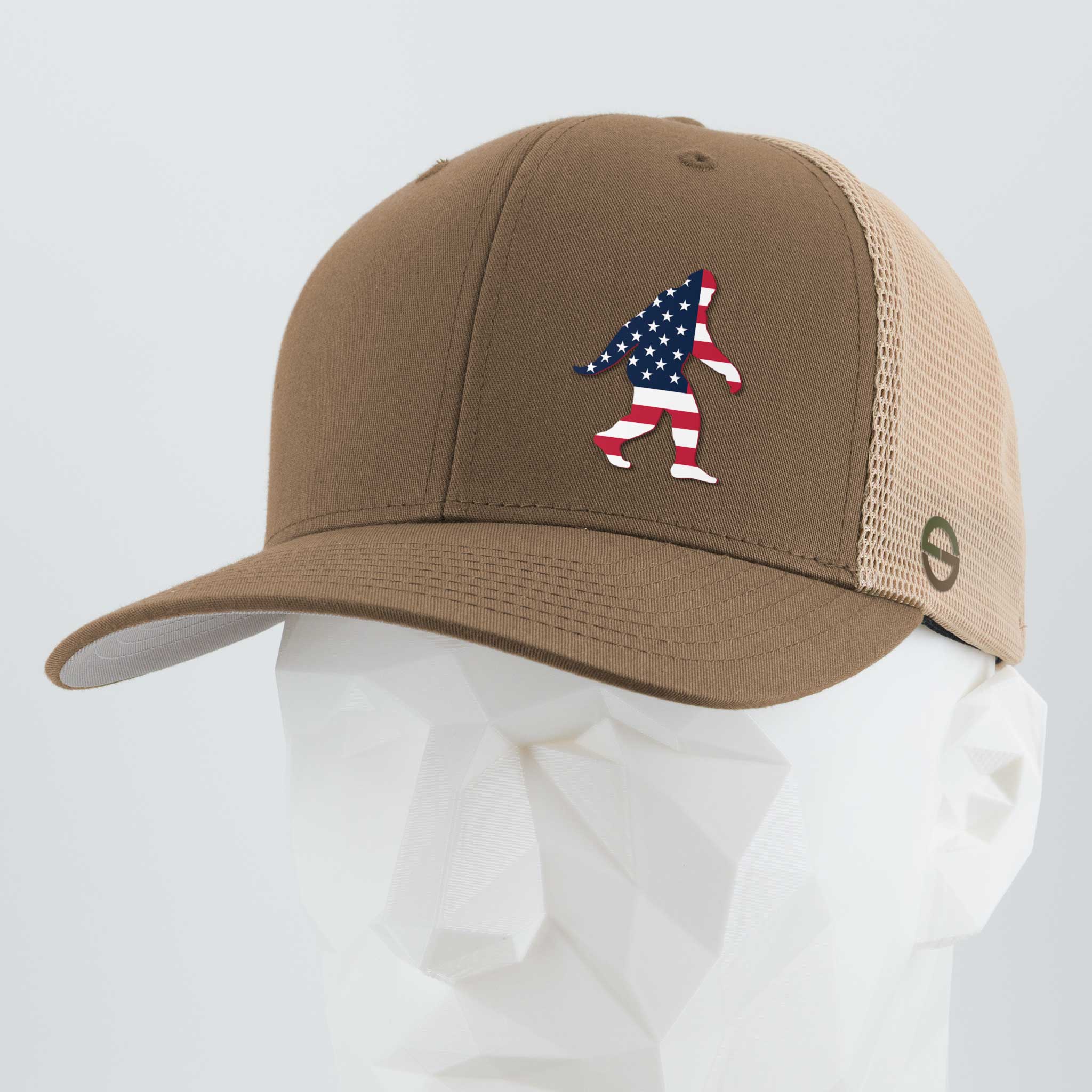 The Eternal Glyph Patriotic Bigfoot (Silicone) Flexfit® Mesh Trucker Cap features a tan and beige design with an American flag-patterned Bigfoot silhouette, displayed here on a white mannequin head.