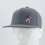 The Eternal Glyph Patriotic Bigfoot (Silicone) Flexfit® Flat Bill Premium Fitted Cap is a gray snapback hat with an American flag Bigfoot silhouette, shown on a white geometric mannequin—a tribute to Sasquatch legends and U.S. pride.