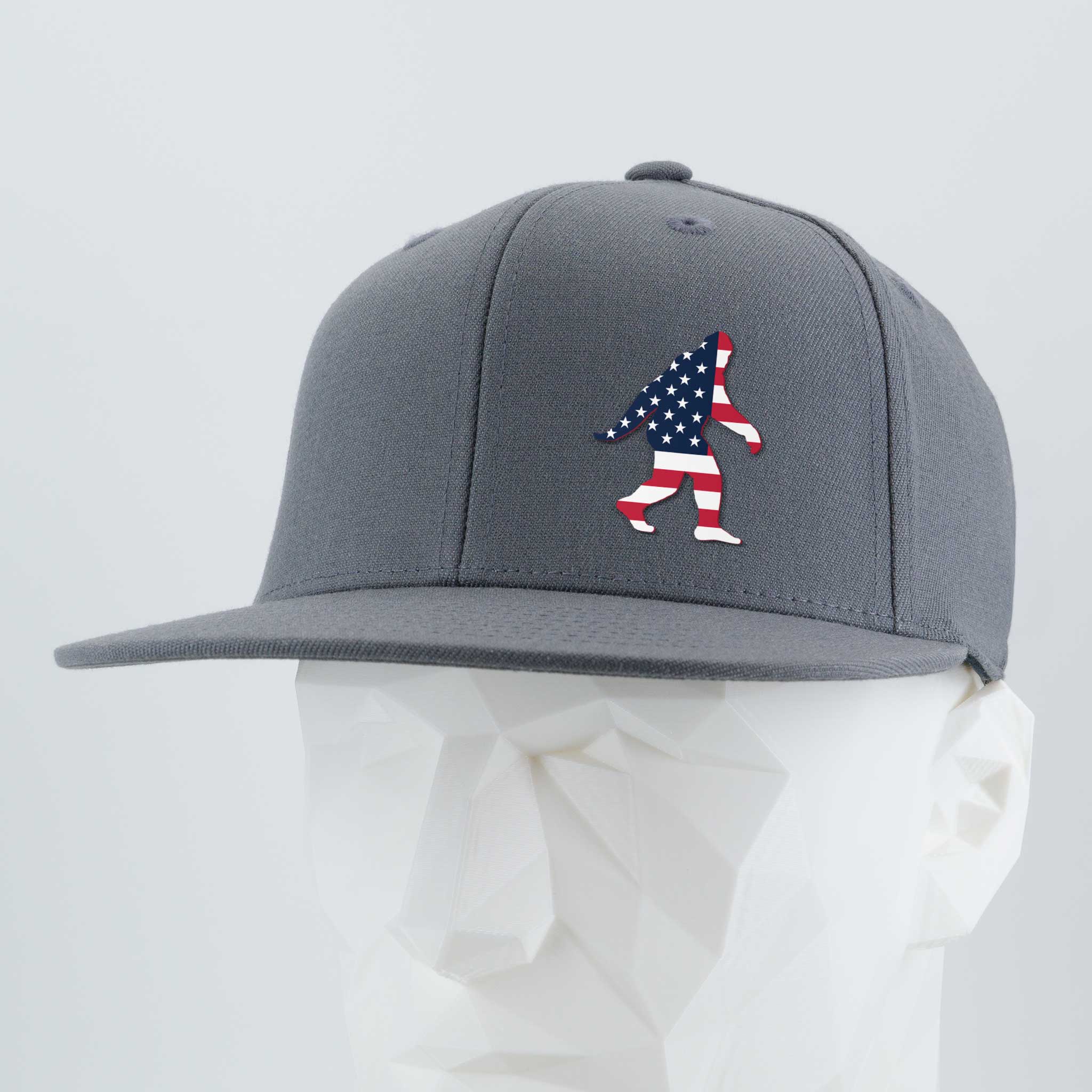 The Eternal Glyph Patriotic Bigfoot (Silicone) Flexfit® Flat Bill Premium Fitted Cap is a gray snapback hat with an American flag Bigfoot silhouette, shown on a white geometric mannequin—a tribute to Sasquatch legends and U.S. pride.