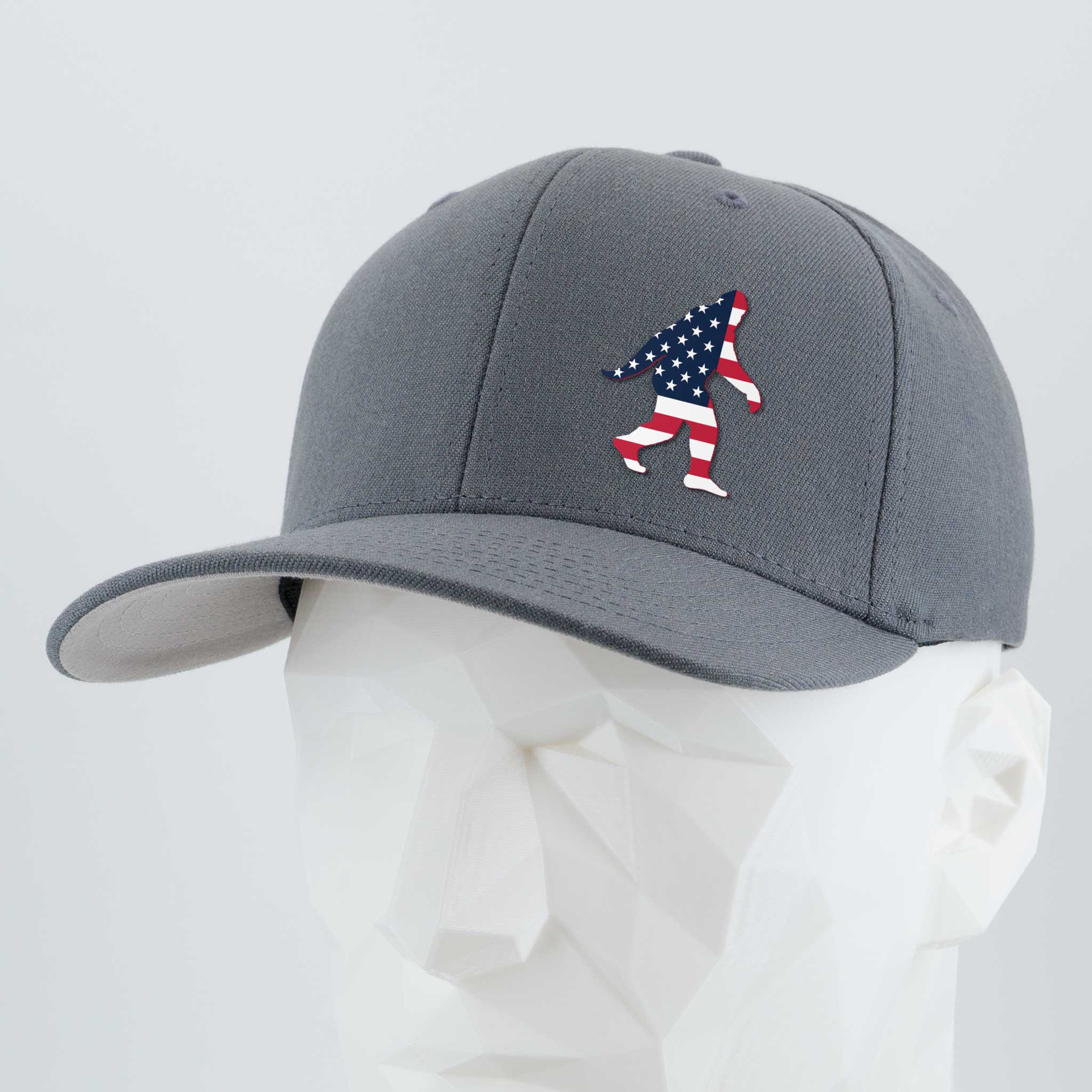 The Eternal Glyph Patriotic Bigfoot (Silicone) Flexfit® Premium Wool Blend Cap in gray features an American flag Bigfoot silhouette on the front, shown on a white geometric mannequin head.