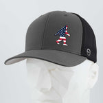 The Eternal Glyph Patriotic Bigfoot (Silicone) Flexfit® Mesh Trucker Cap in dark gray and black features a small American flag Sasquatch silhouette on the front, shown here on a white geometric mannequin head.
