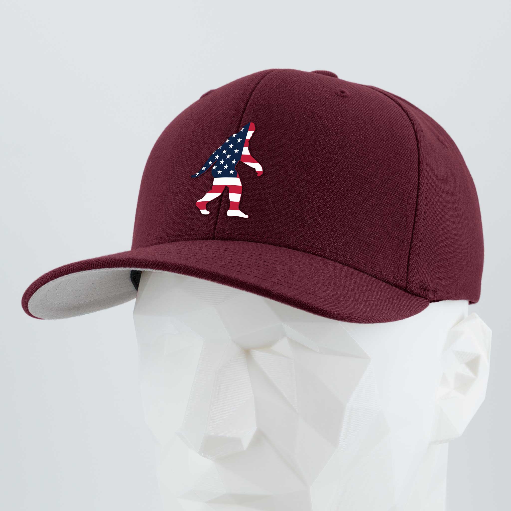 A maroon Flexfit® Premium Wool Blend Cap by Eternal Glyph, shown on a white mannequin head, features a silicone American flag Bigfoot silhouette on the front—a tribute to wilderness folklore.
