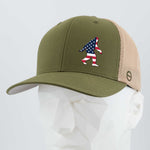 The Eternal Glyph Patriotic Bigfoot (Silicone) Flexfit® Mesh Trucker Cap in olive green and beige features an American flag Sasquatch patch—ideal for wilderness folklore fans and patriotic Bigfoot enthusiasts. Displayed on a white mannequin head.