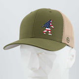 The Eternal Glyph Patriotic Bigfoot (Silicone) Flexfit® Mesh Trucker Cap in olive green and beige features an American flag Sasquatch patch—ideal for wilderness folklore fans and patriotic Bigfoot enthusiasts. Displayed on a white mannequin head.