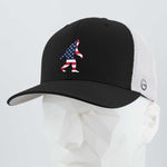 The Eternal Glyph Patriotic Bigfoot (Silicone) Flexfit® Mesh Trucker Cap in Black/White features a bold American flag Bigfoot silhouette, shown on a white faceted mannequin—ideal for wilderness folklore fans.