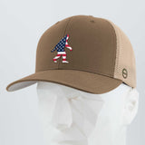The Eternal Glyph Patriotic Bigfoot (Silicone) - Flexfit® Mesh Trucker Cap in Coyote/Khaki features an American flag Bigfoot silhouette—ideal for wilderness folklore fans. Displayed on a white geometric mannequin head.