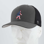 The Eternal Glyph Patriotic Bigfoot (Silicone) Flexfit® Mesh Trucker Cap in dark gray/black features a bold American flag Bigfoot design on the front, displayed on a white mannequin—ideal for fans of legendary wilderness adventures.