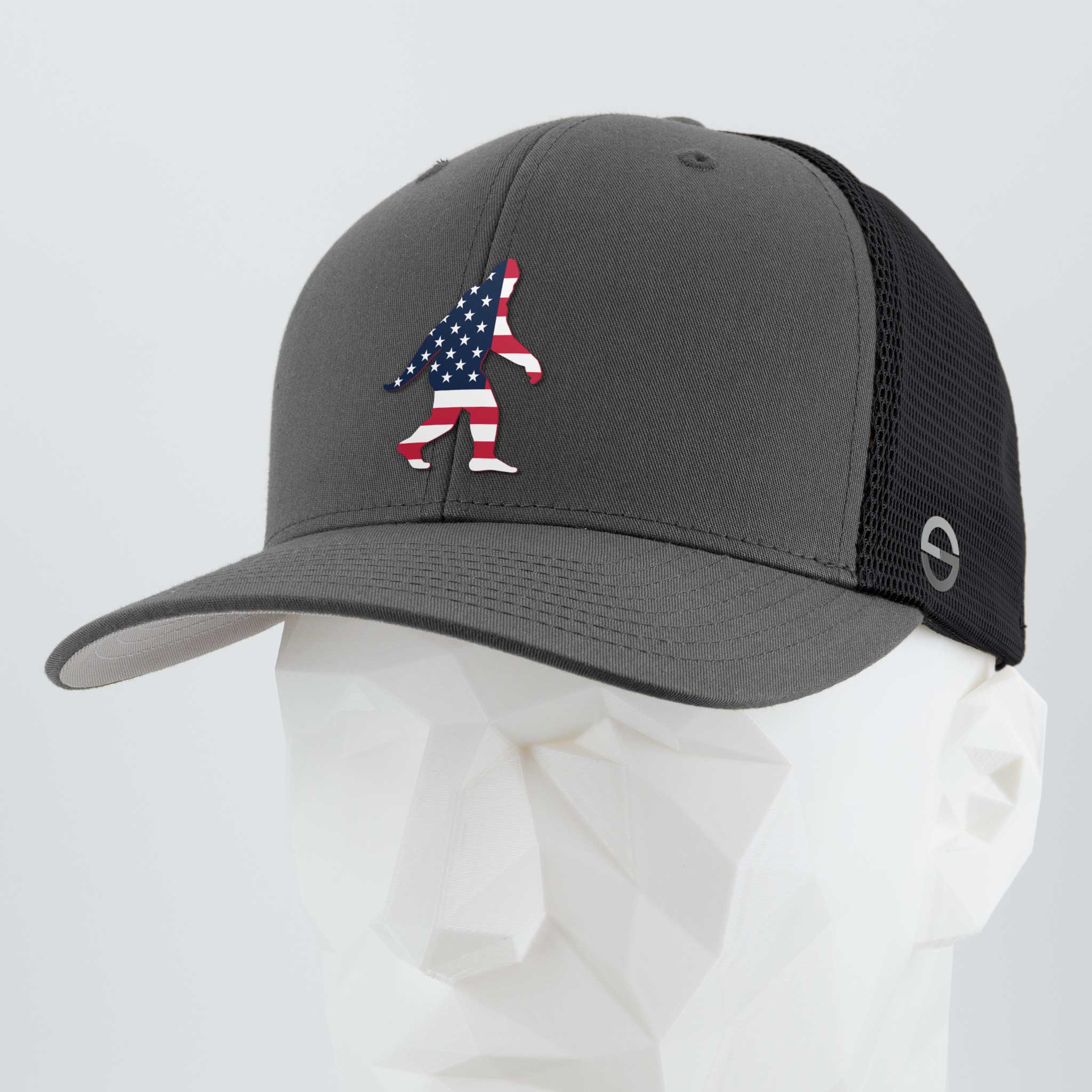 The Eternal Glyph Patriotic Bigfoot (Silicone) Flexfit® Mesh Trucker Cap in dark gray/black features a bold American flag Bigfoot design on the front, displayed on a white mannequin—ideal for fans of legendary wilderness adventures.