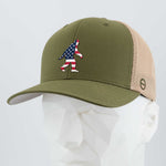 The Eternal Glyph Patriotic Bigfoot (Silicone) Flexfit® Mesh Trucker Cap in Olive/Khaki features an American flag Bigfoot silhouette, blending wilderness folklore with unique style—displayed on a white mannequin.