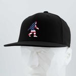 The Eternal Glyph Patriotic Bigfoot (Silicone) Flexfit® Flat Bill Premium Fitted Cap features a black design with an embroidered American flag Bigfoot, displayed on a white geometric mannequin against a gray background.