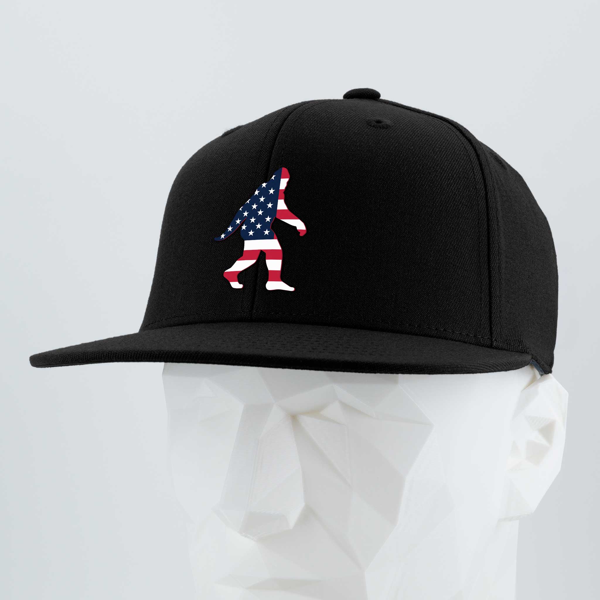 The Eternal Glyph Patriotic Bigfoot (Silicone) Flexfit® Flat Bill Premium Fitted Cap features a black design with an embroidered American flag Bigfoot, displayed on a white geometric mannequin against a gray background.