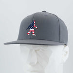 The Eternal Glyph Patriotic Bigfoot (Silicone) Flexfit® Flat Bill Premium Fitted Cap showcases an American flag Bigfoot silhouette on gray—ideal for wilderness folklore fans. Displayed on a faceted white mannequin.
