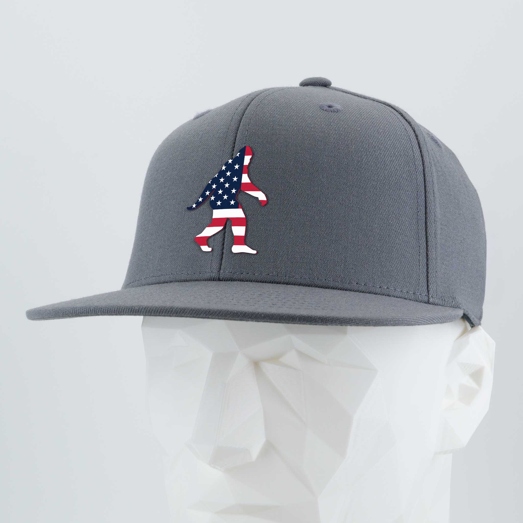 The Eternal Glyph Patriotic Bigfoot (Silicone) Flexfit® Flat Bill Premium Fitted Cap showcases an American flag Bigfoot silhouette on gray—ideal for wilderness folklore fans. Displayed on a faceted white mannequin.