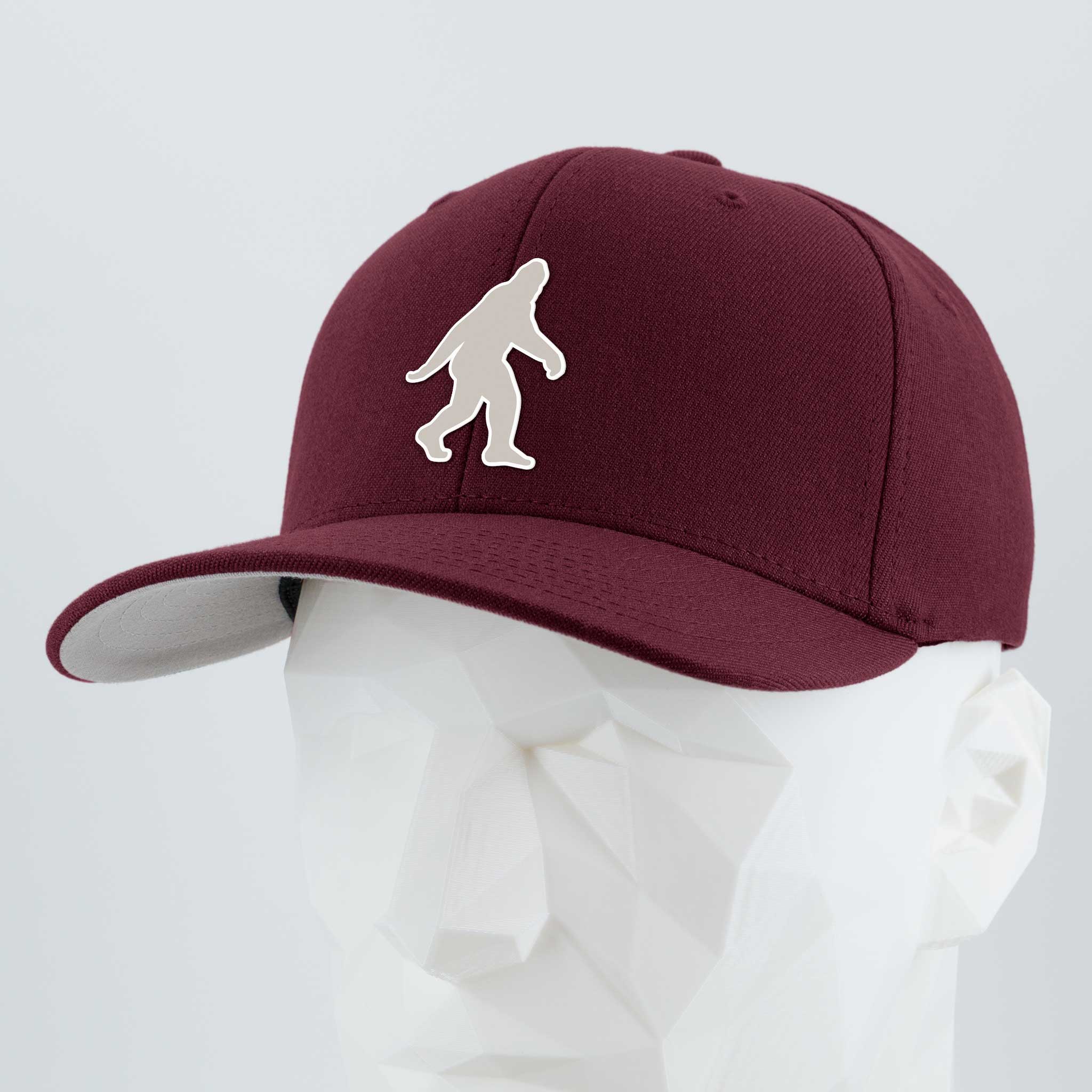 The Eternal Glyph Bigfoot Silhouette Albino (Silicone) Flexfit® Premium Wool Blend Cap in maroon, featuring a white Bigfoot design inspired by Pacific Northwest folklore, is displayed on a white geometric mannequin head against a plain background.