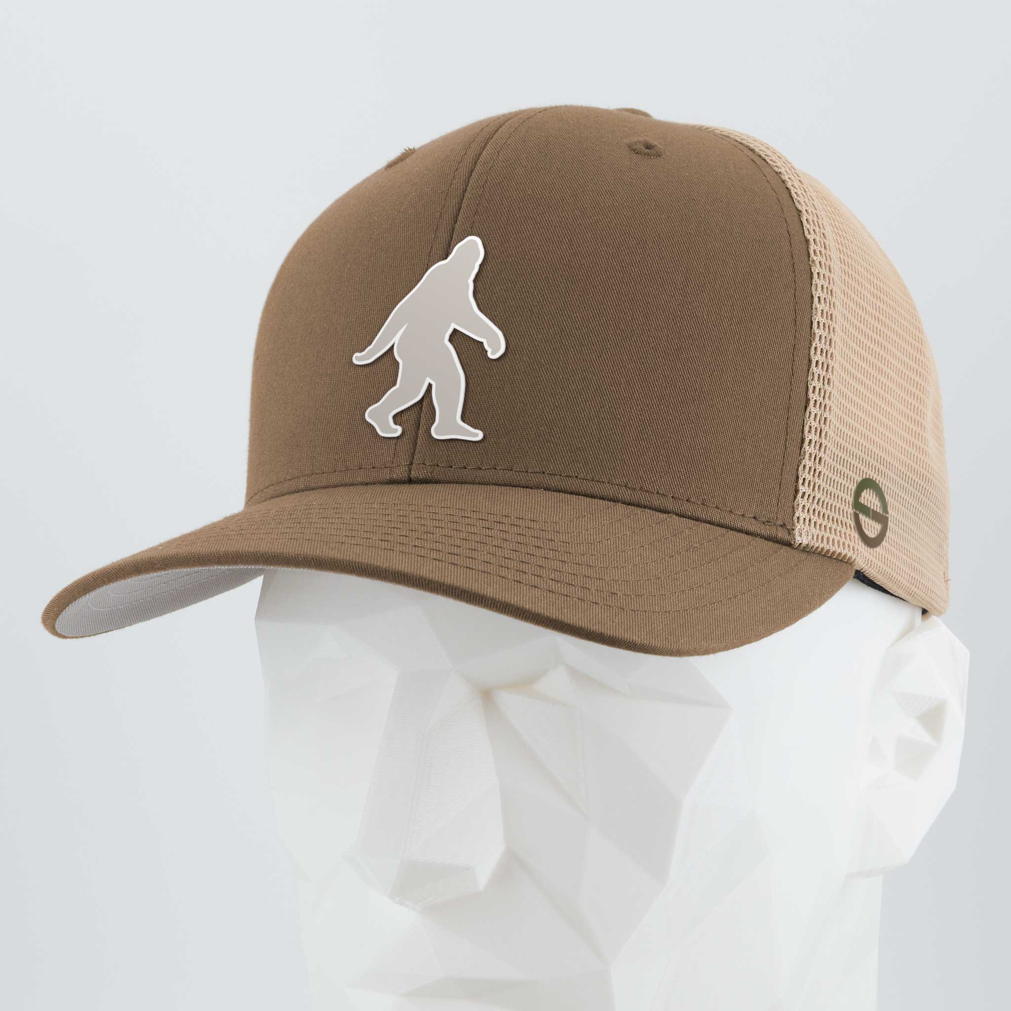 The Eternal Glyph Bigfoot Silhouette Albino (Silicone) Flexfit® Mesh Trucker Cap in coyote/khaki features a bold Bigfoot design, inspired by Pacific Northwest folklore, shown on a white geometric mannequin head.