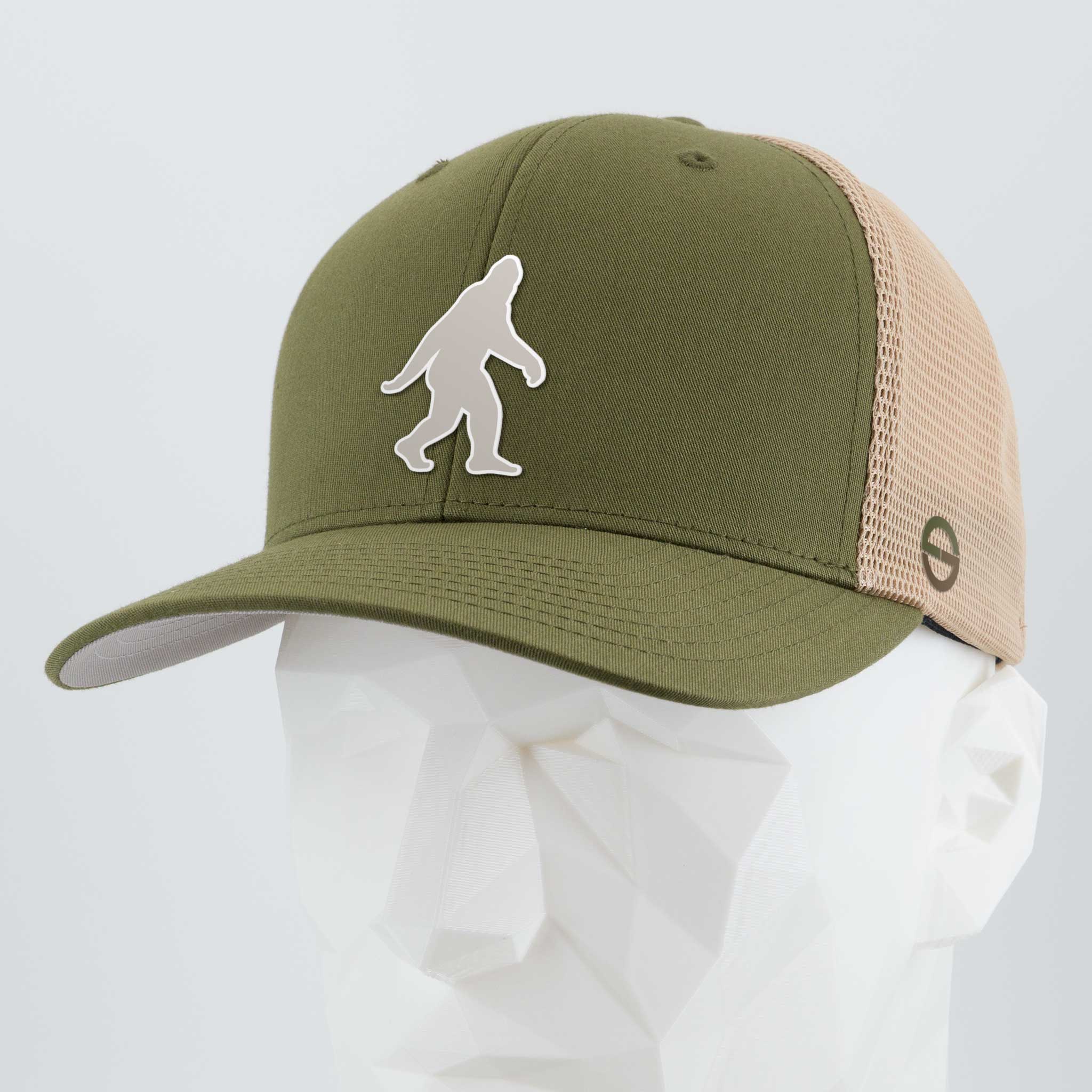 The Eternal Glyph Flexfit® Mesh Trucker Cap in Olive/Khaki features a white Bigfoot Silhouette Albino (Silicone) logo, inspired by Pacific Northwest folklore, shown here on a white geometric mannequin head.